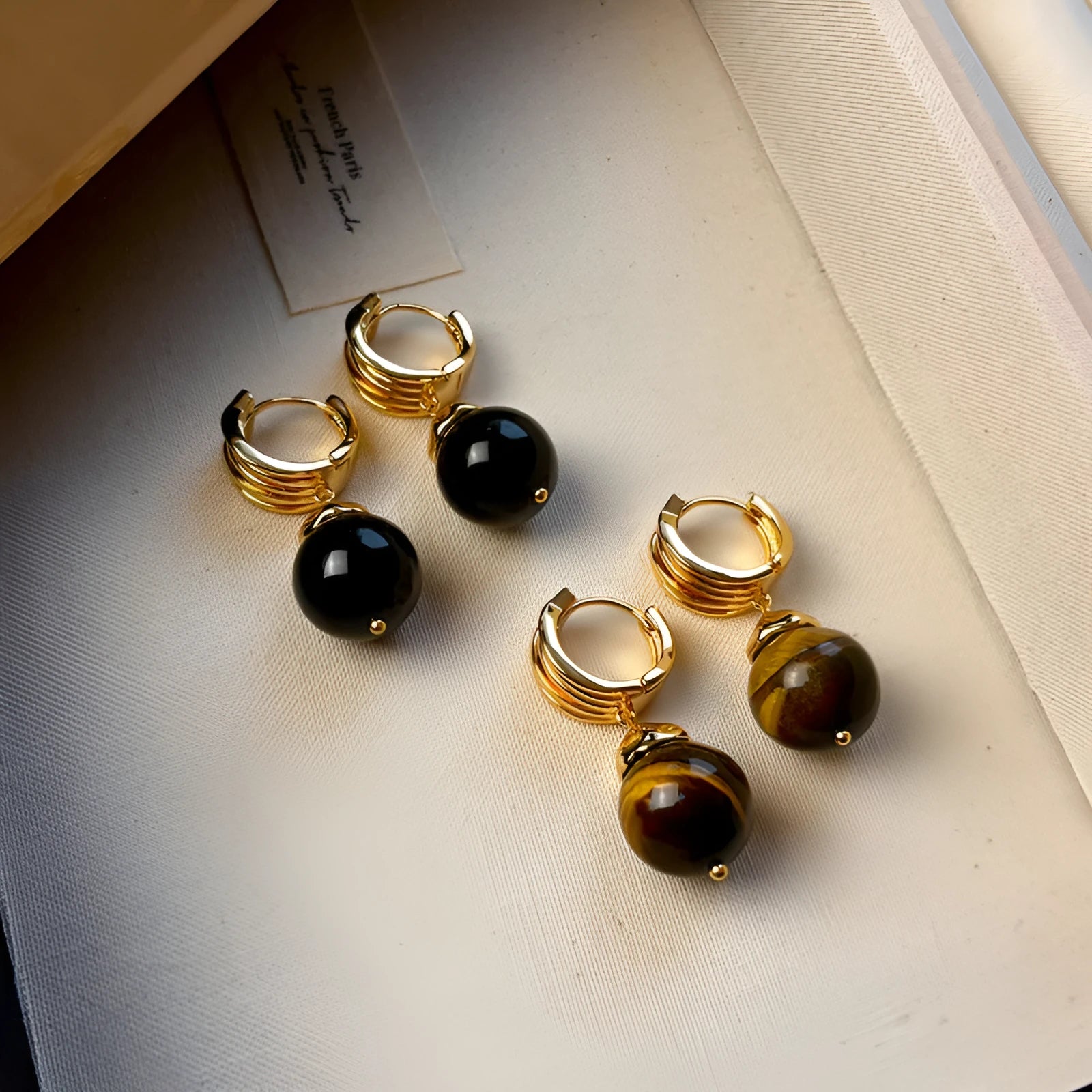 A pair of geometric tigereye stone earrings with 18K gold plating, showing natural brown bands, on a grey background.