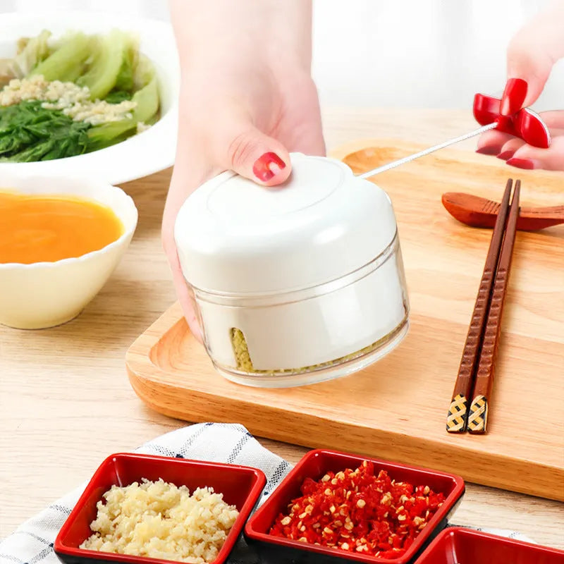 White manual food chopper with stainless steel blades on a magnetic holder, shown with chopped vegetables.