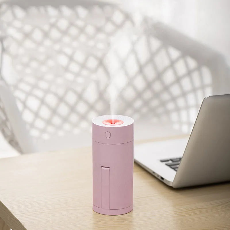 A compact white folding bracket USB humidifier on a desktop next to a laptop, showing its mist output.