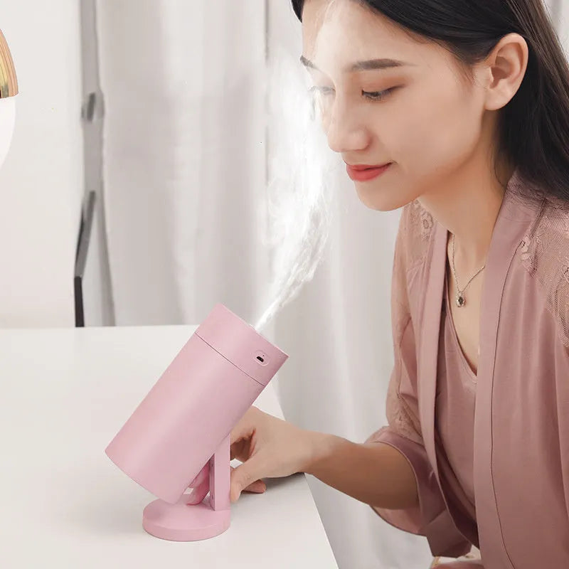 A compact white folding bracket USB humidifier on a desktop next to a laptop, showing its mist output.