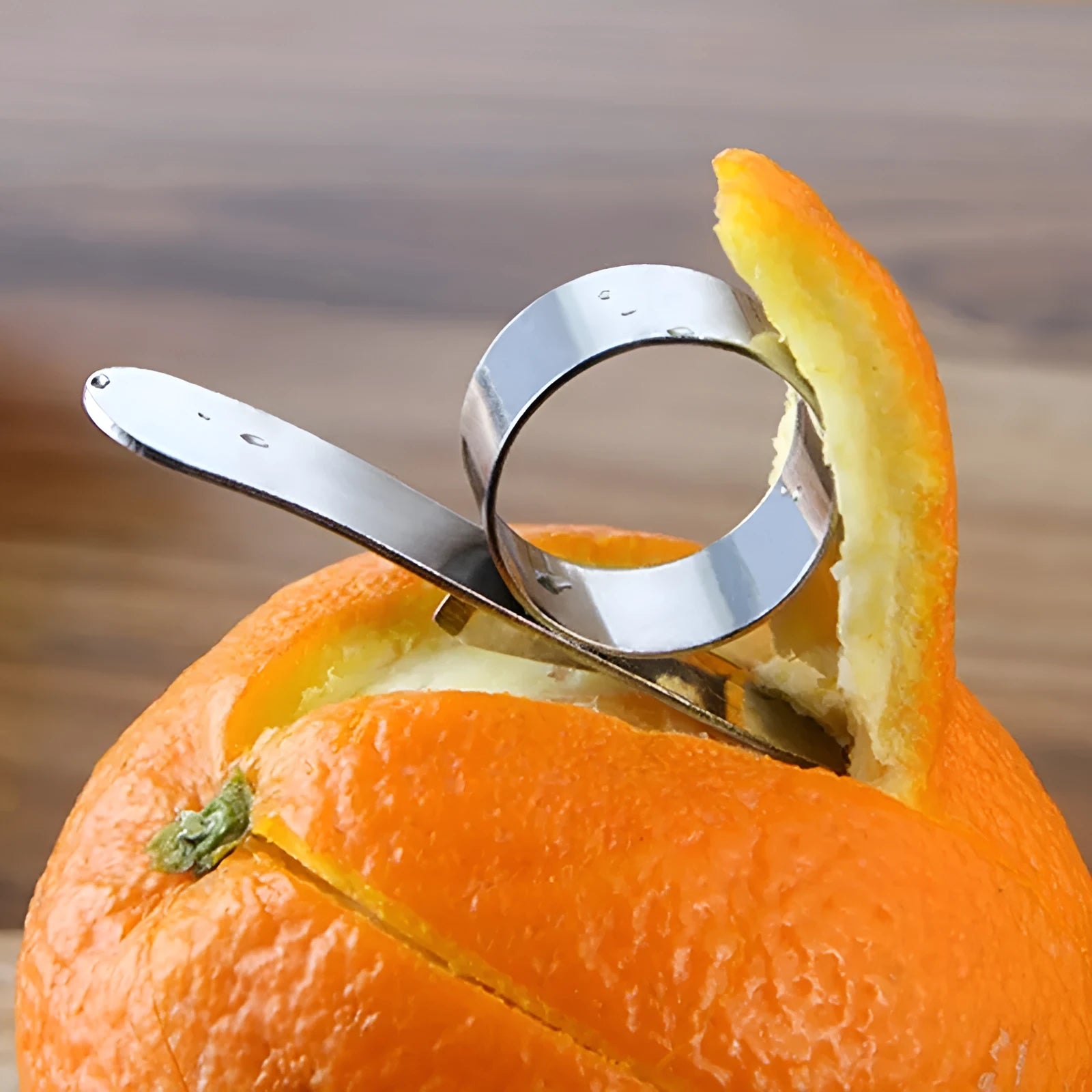 A silver stainless steel finger-ring style citrus peeler for oranges and pomelo on a kitchen surface.
