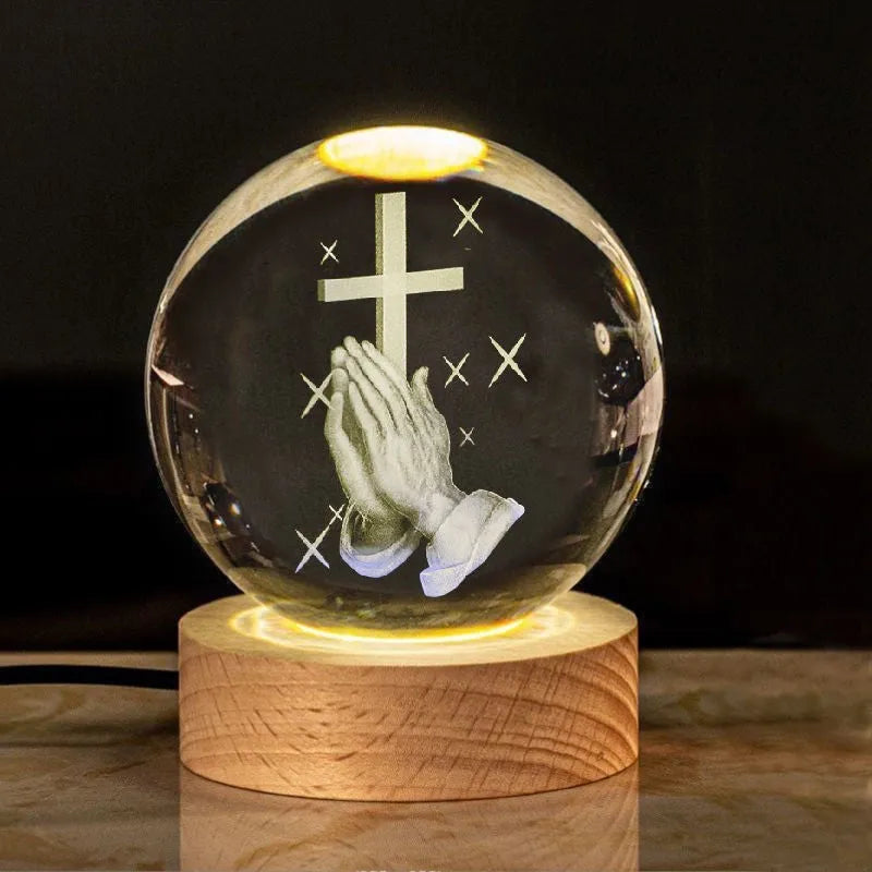 White crystal sphere on wooden base with warm glow, showing religious symbols including cross and prayer hands