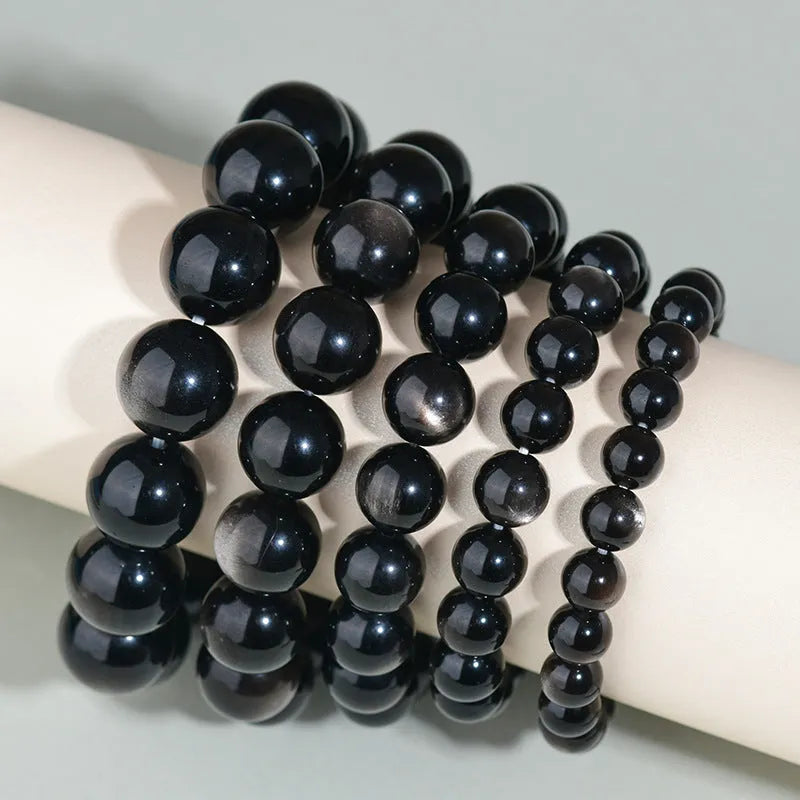 Hand-woven obsidian bracelet with round beads in adjustable sizes from 8mm to 16mm diameter, showing six colour options including black stone and rainbow eye obsidian.