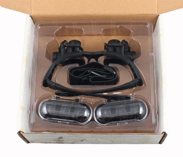 Black hands-free magnifier glasses with LED lights and four pairs of interchangeable magnification lenses on a workbench.