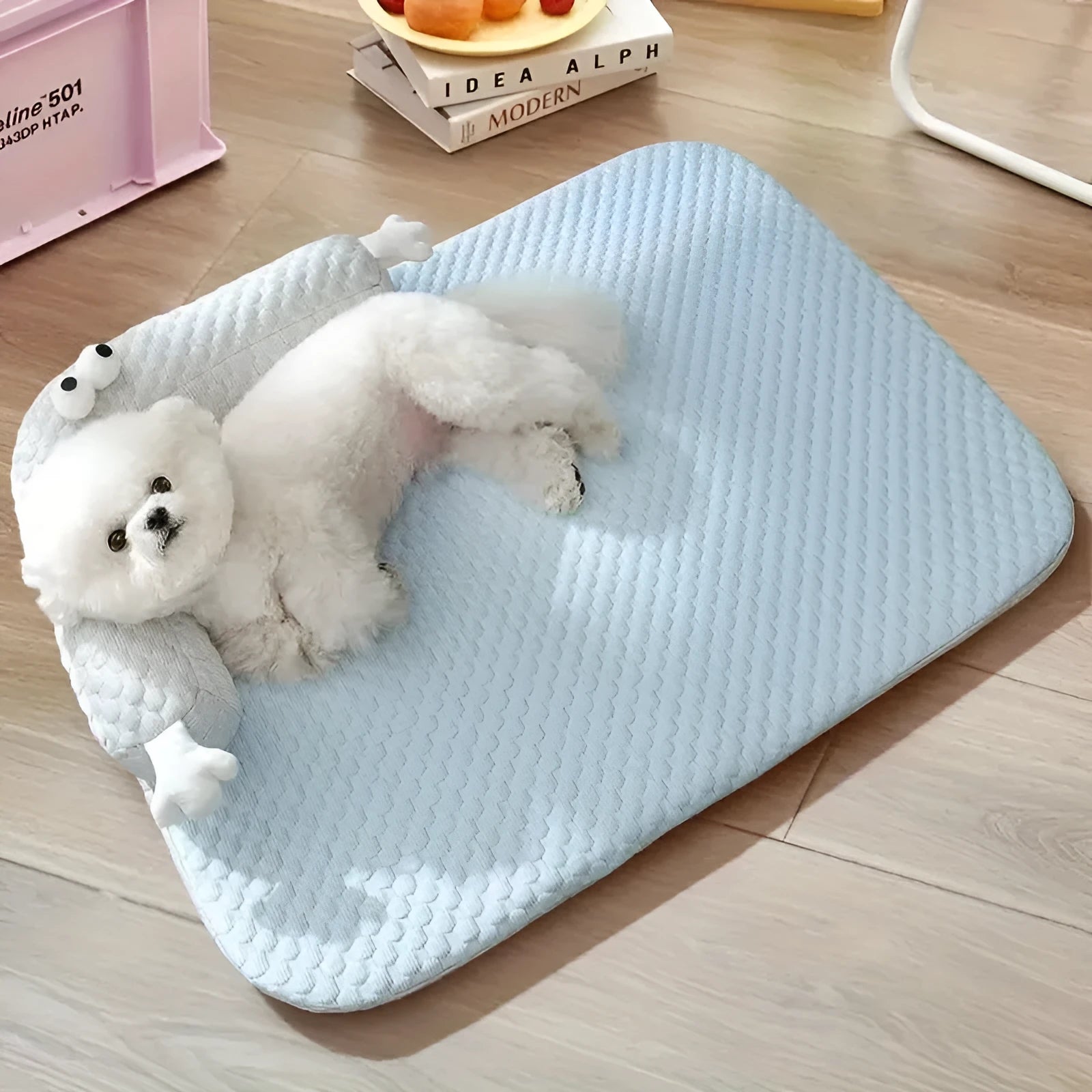 Blue pet bed cushion with big-eyed monster design on a wooden floor.