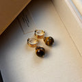 A pair of geometric tigereye stone earrings with 18K gold plating, showing natural brown bands, on a grey background.