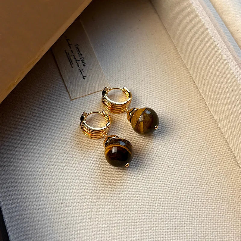 A pair of geometric tigereye stone earrings with 18K gold plating, showing natural brown bands, on a grey background.