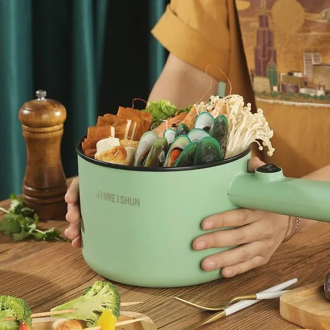 White and green mini electric hot pot cooker with stainless steel body and cooking accessories on a kitchen counter.