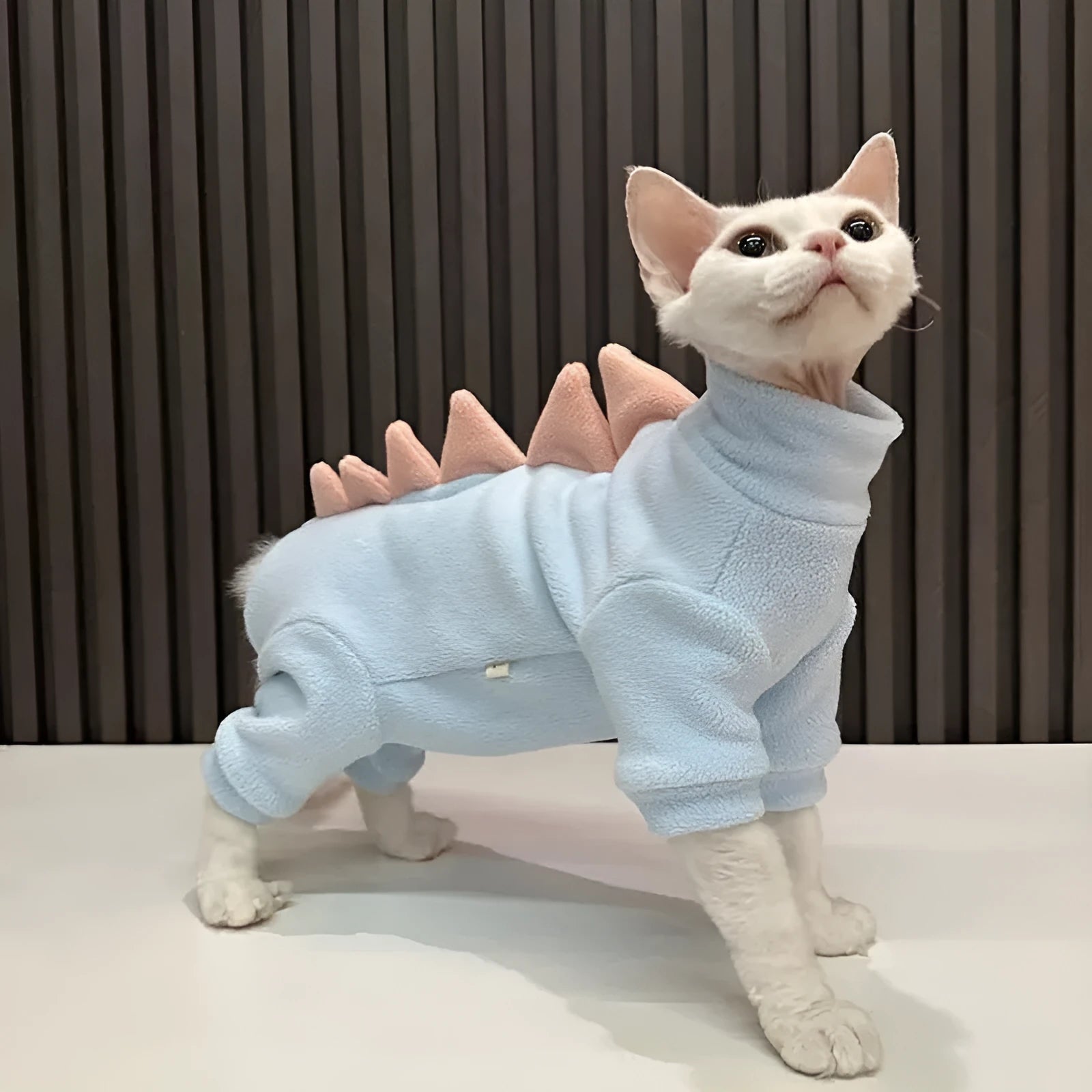 Sphynx cat wearing green dinosaur costume with tail and spikes, showing full-body fleece outfit