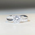 Geometric silver moissanite ring with twisted arm design and four-claw setting, shown in white diamond colour.