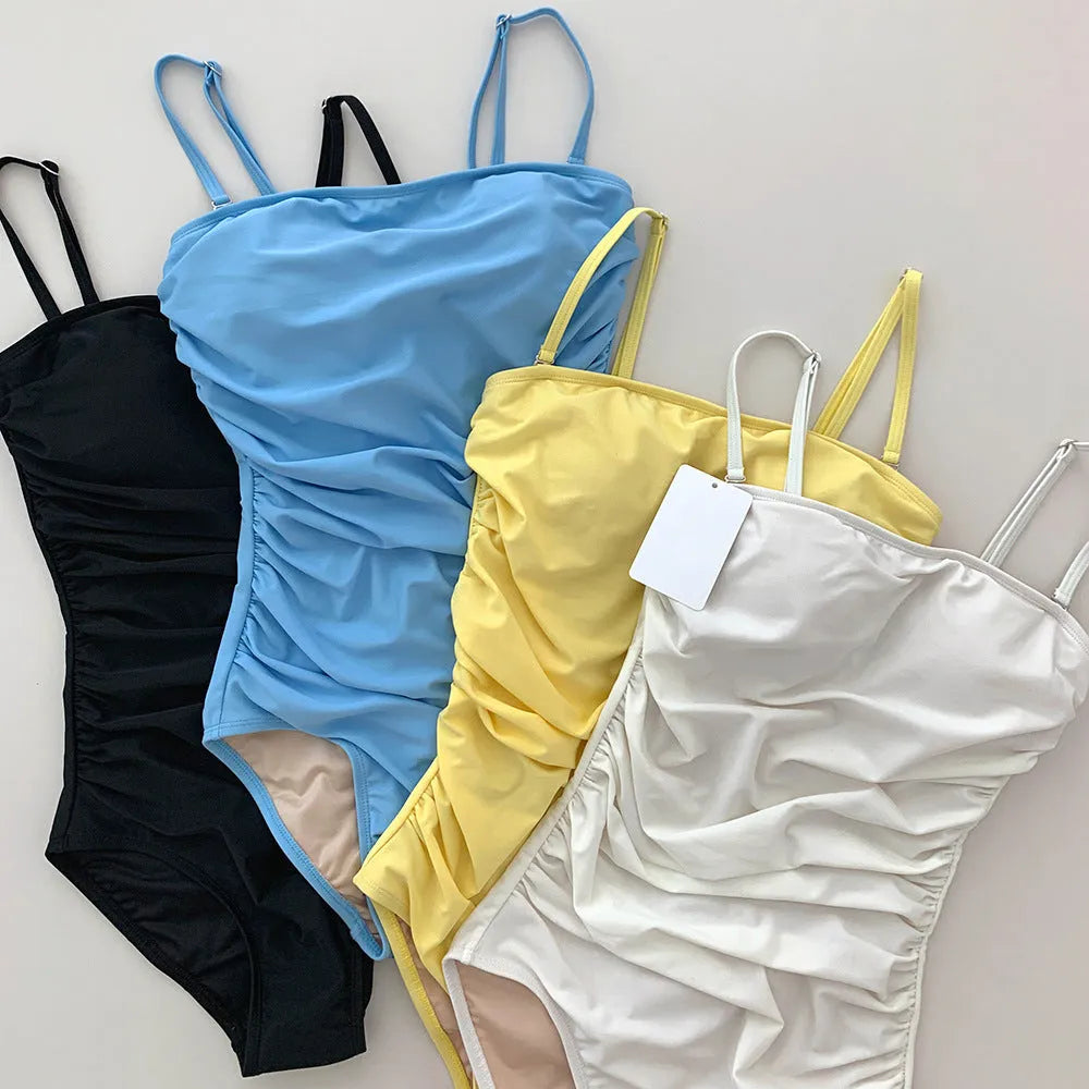 Sling style one-piece swimsuit with belly pleats in black, blue, white, or yellow on a model