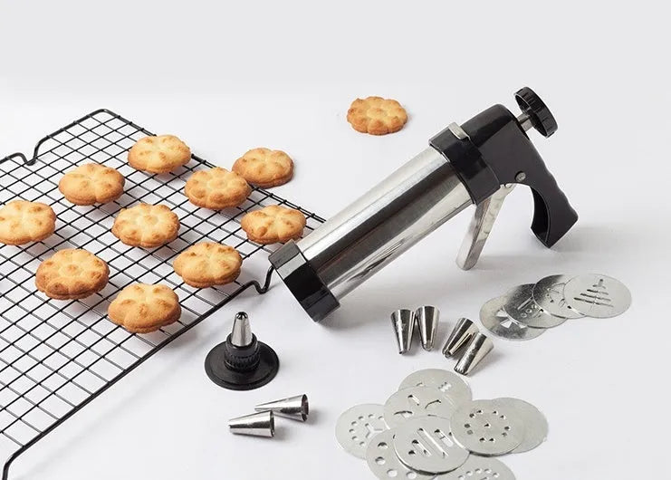 Stainless steel biscuit press gun with assorted cookie moulds and piping nozzles on a baking tray.