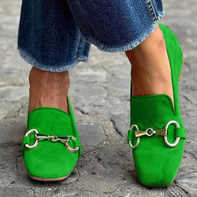 Green suede loafers with metal fastener, low heel, and rubber soles on a wooden floor