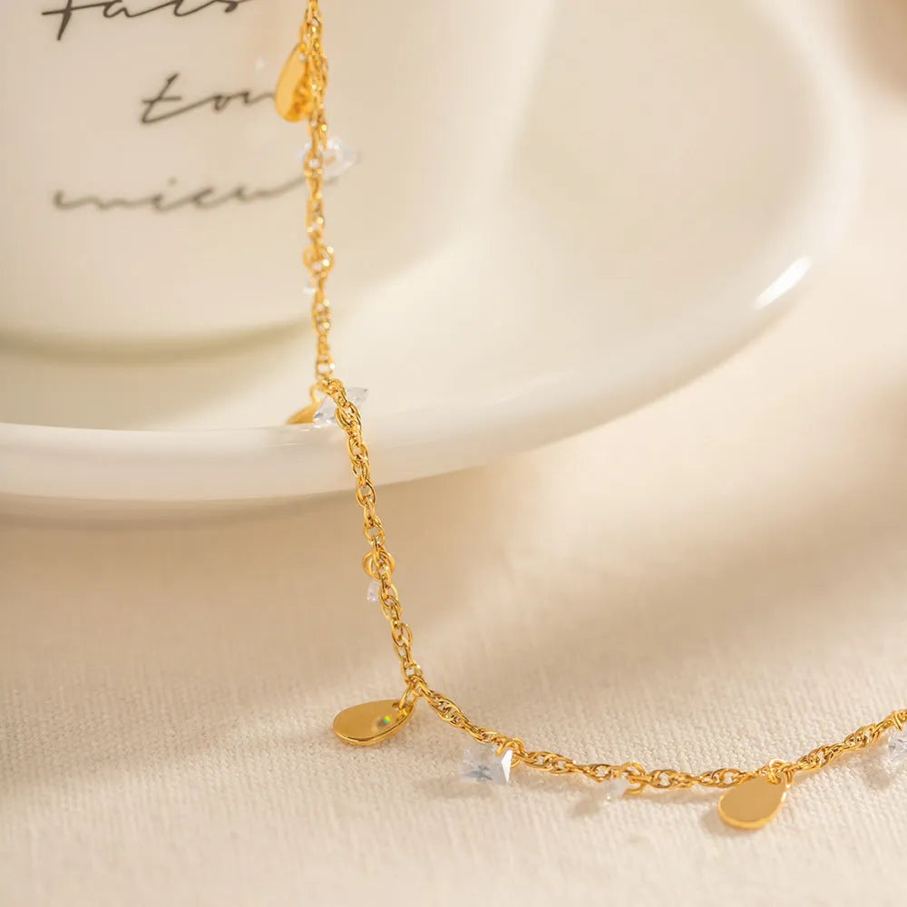 Close-up of a stainless steel anklet with a geometric water drop pendant and dangling tassel charm on an ankle.