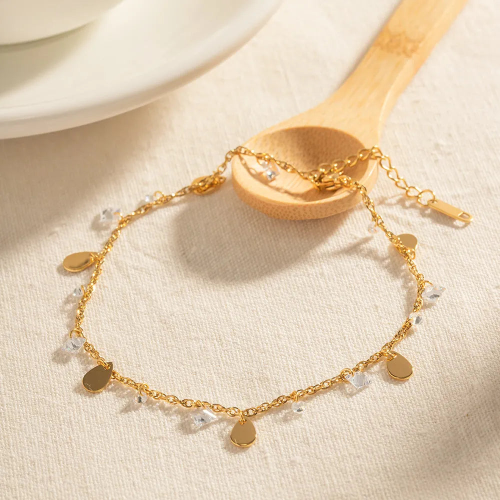 Close-up of a stainless steel anklet with a geometric water drop pendant and dangling tassel charm on an ankle.
