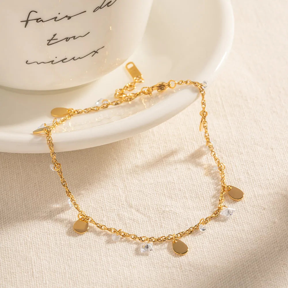 Close-up of a stainless steel anklet with a geometric water drop pendant and dangling tassel charm on an ankle.