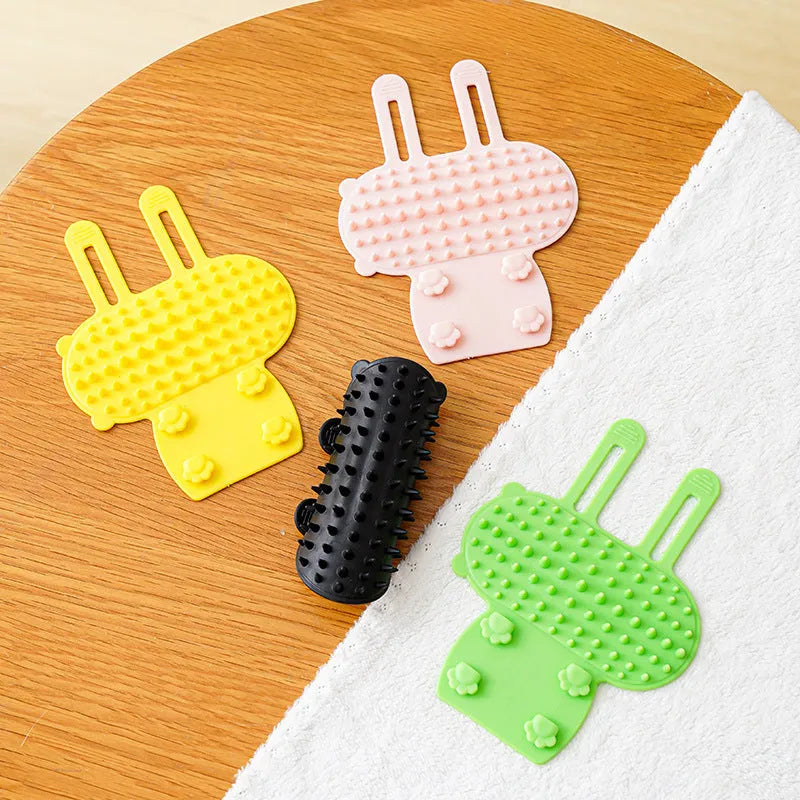Black silicone cat grooming comb with adjustable strap attached to a table leg, showing rounded teeth for hair removal