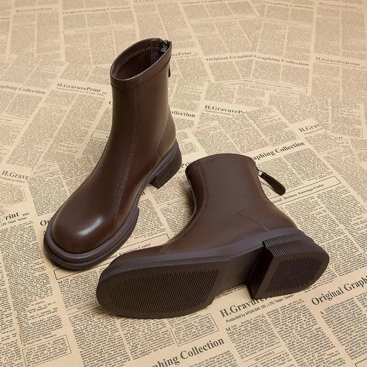 Women's black ankle boots with a thick flat heel, round toe, and rear zip fastener on a wooden floor.