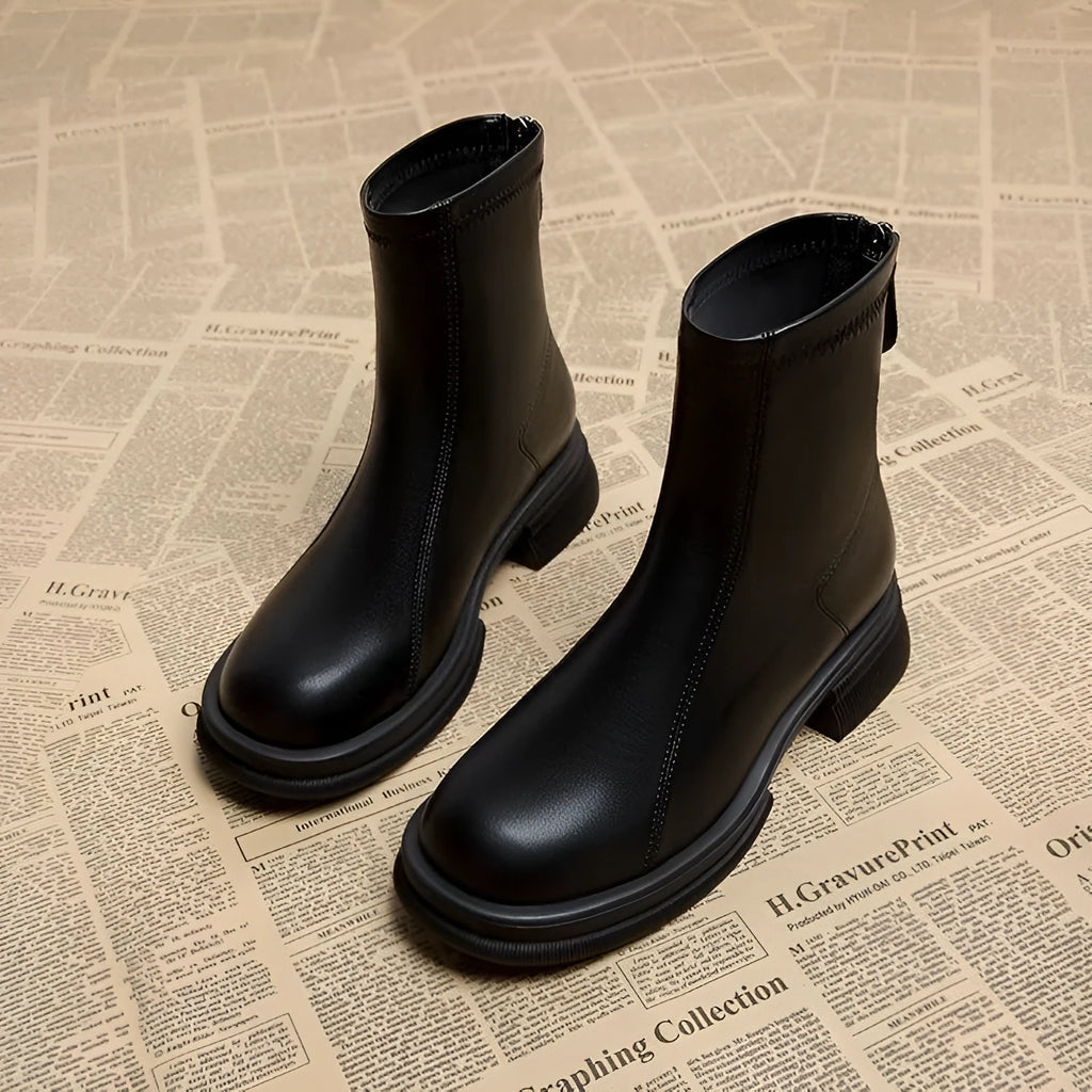 Women's black ankle boots with a thick flat heel, round toe, and rear zip fastener on a wooden floor.